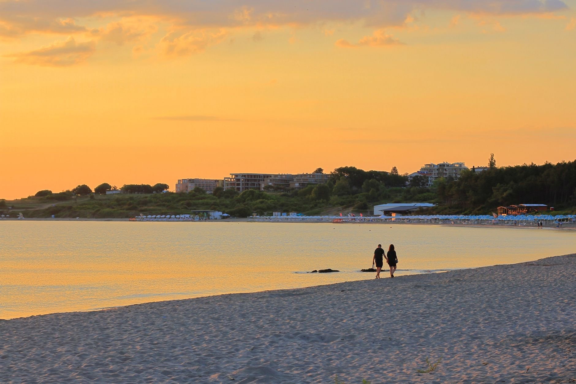 Couple walking on a sandy beach during a beautiful sunset, with a calm sea in the distance.
